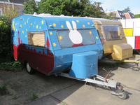 833739 Afbeelding van een caravan beschilderd met een stadssilhouet en een nijntje, op het parkeerterrein bij een loods ...